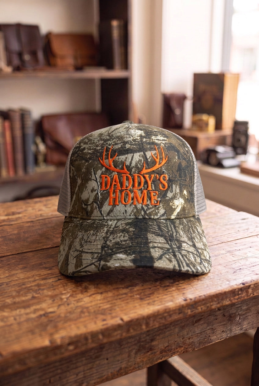 DADDY'S HOME embroidered camo trucker hat, front view on rustic wood table